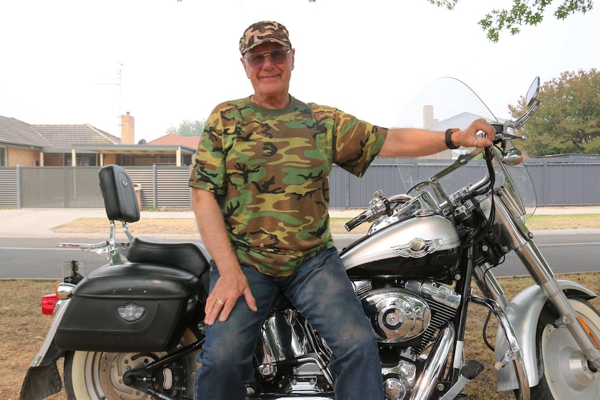 A man in a camouflage shirt and hat sits smiling happily on a Harley Davidson motorbike.