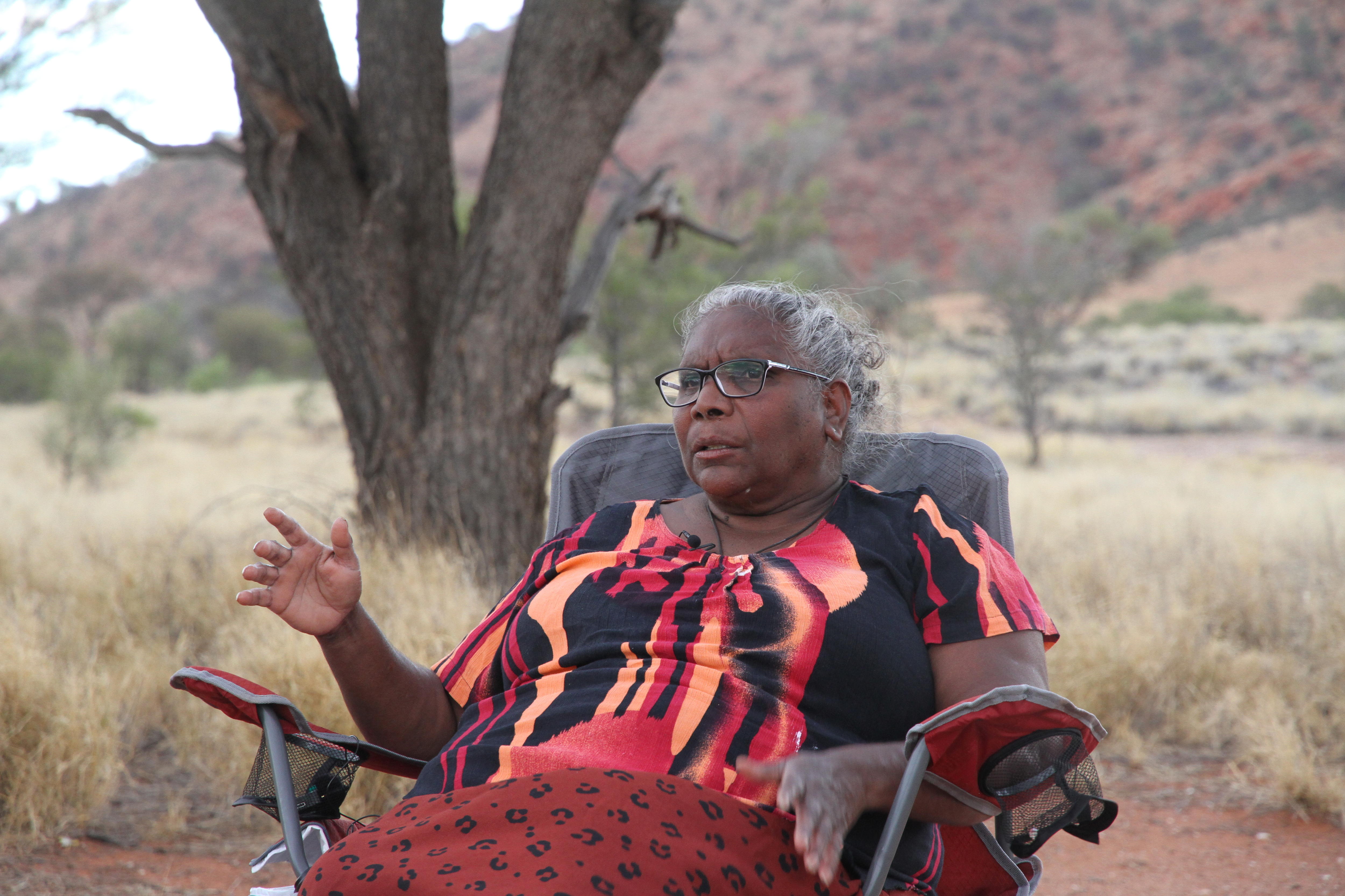She sits in a camp chair wearing colourful red and orange clothes