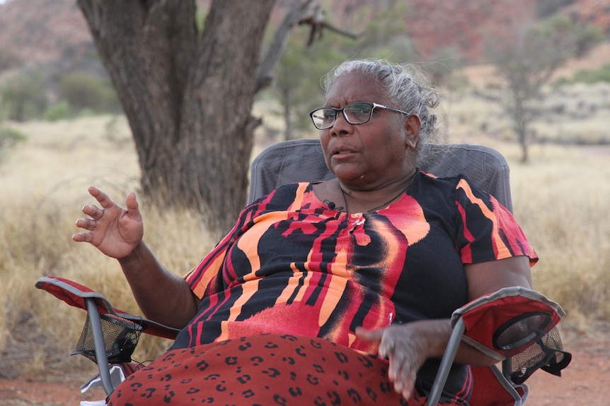 She sits in a camp chair wearing colourful red and orange clothes