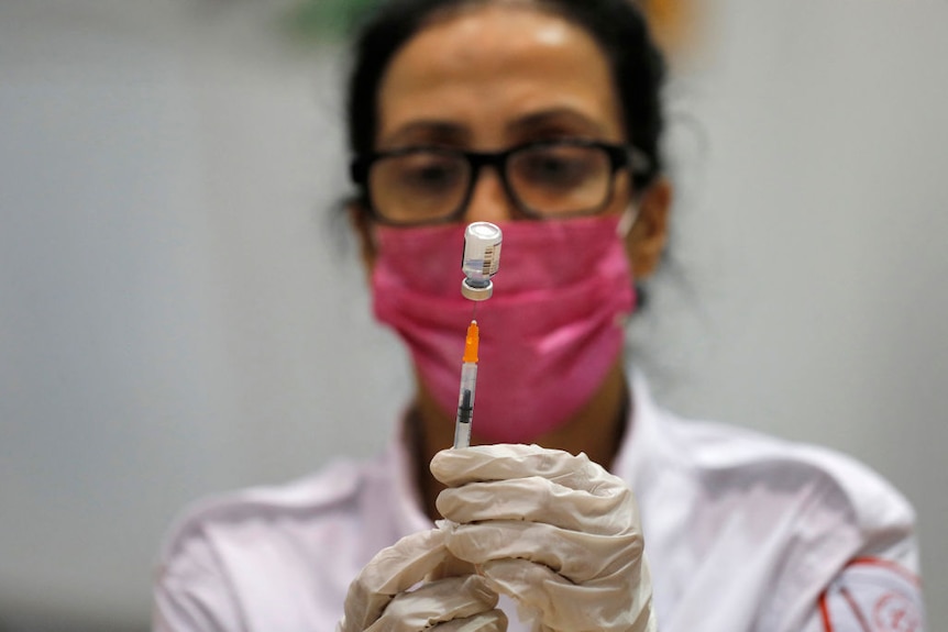 Paramedic wearing glasses and pink face mask holds up vaccine in front of her face,