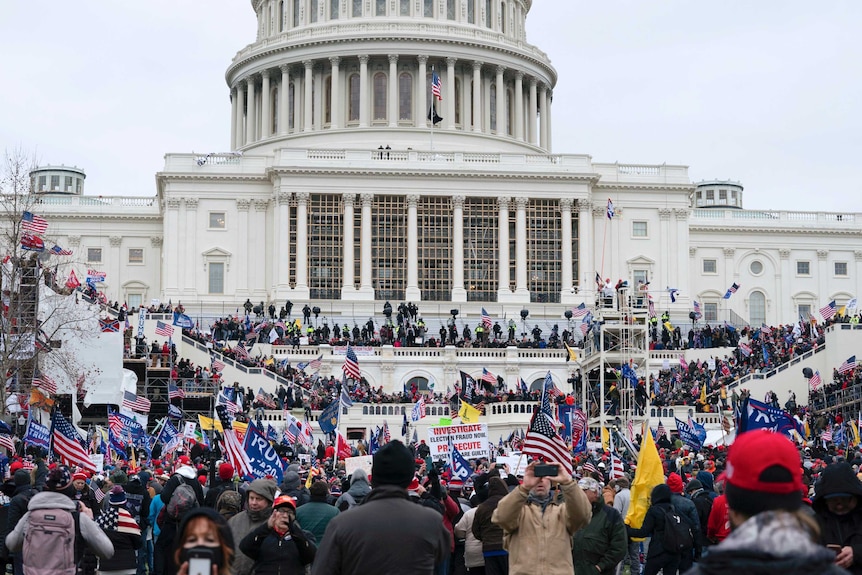 A large crowd of people gather outside the US Capitol building. Many are waving US flags, or Donald Trump supporter gear