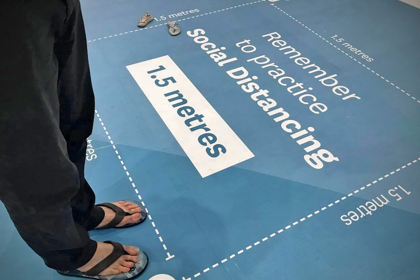 A pair of feet stand near a large sticker on the floor of a shopping centre which shows physical distancing spacing