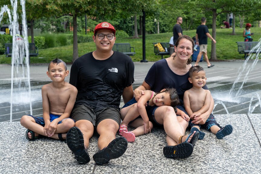 Two parents in black T-shirts and sandals site with their three small children who are wearing bathers at a waterpark
