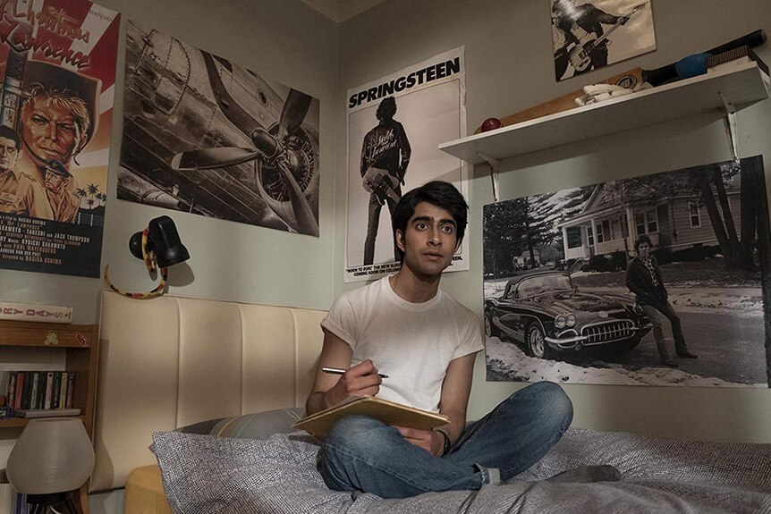 Viveik Kalra wears white t-shirt and blue jeans and is seated on bed in bedroom decorated with Bruce Springsteen posters.