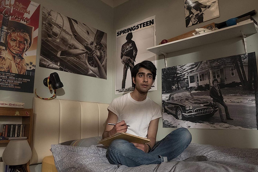 Viveik Kalra wears white t-shirt and blue jeans and is seated on bed in bedroom decorated with Bruce Springsteen posters.