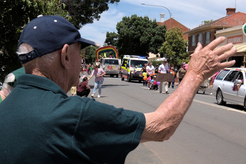 An older man wearing a cap waves to a group of parade participants.