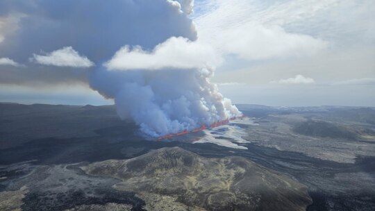 A large plume of smoke billows from a bright orange crack in the ground against a vast landscape