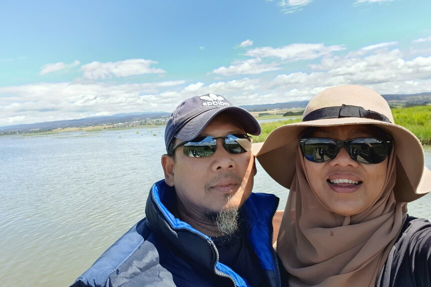A man and woman take a selfie in front of a body of water.