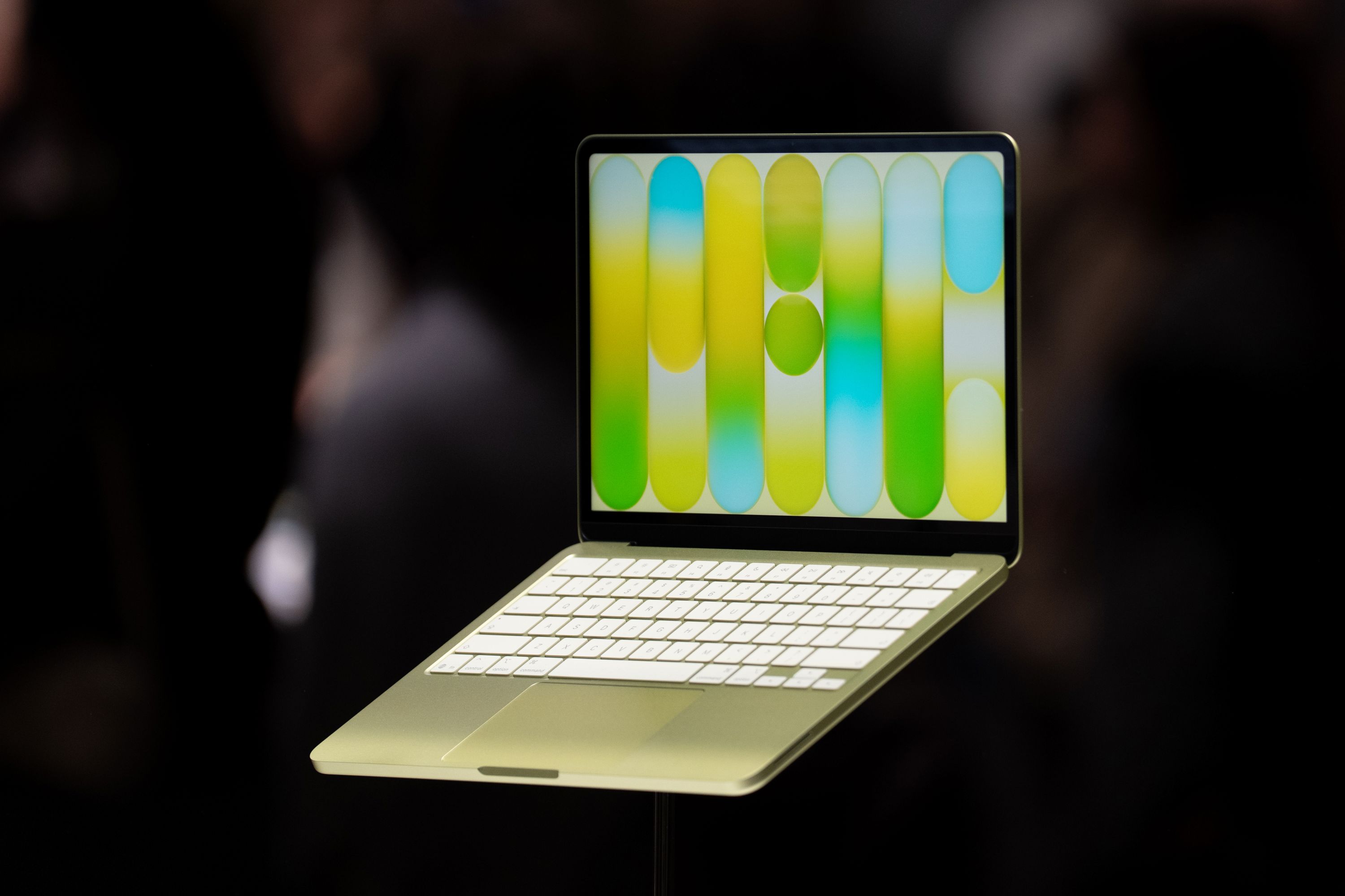 An open silver MacBook Neo laptop with yellow, green and blue graphics on its screen, suspended against a black background.