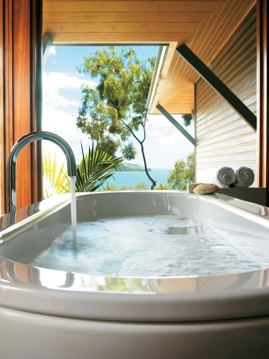 A tap is running to fill up a large bath that looks out through trees onto the ocean.