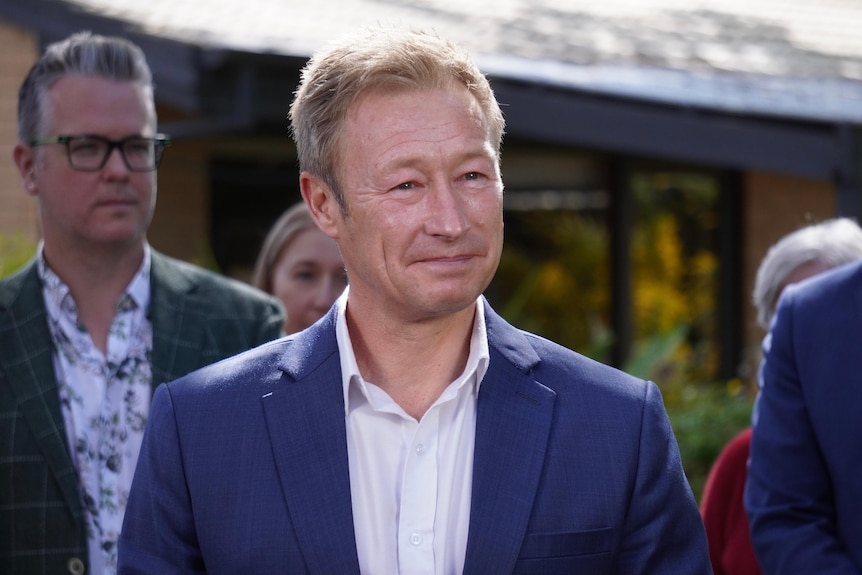 A man wearing a blue suit and light coloured shirt looks slightly off camera, behind him are a group of people