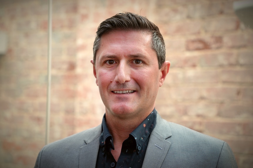 A close up shot of a man in a grey suit standing in front of a well lit brick wall