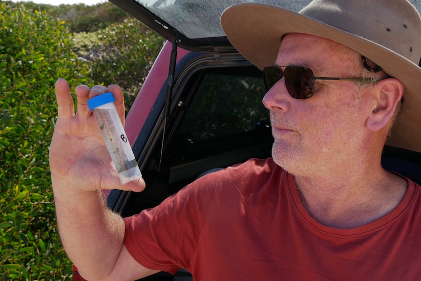 A man in a red shirt and sunglasses looks at a plastic test tube full of water. 