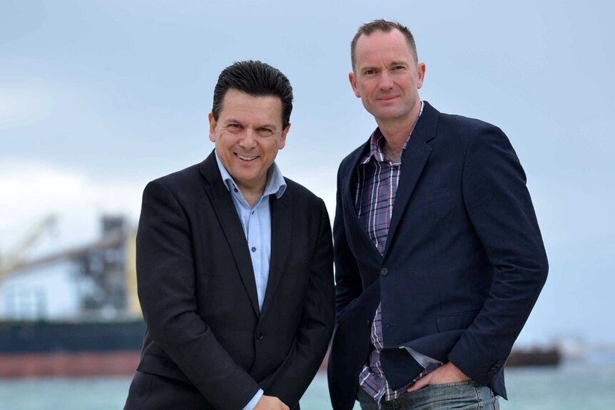 Nick Xenophon with Luke Bolton stand on a beach with a cargo ship docked in the background