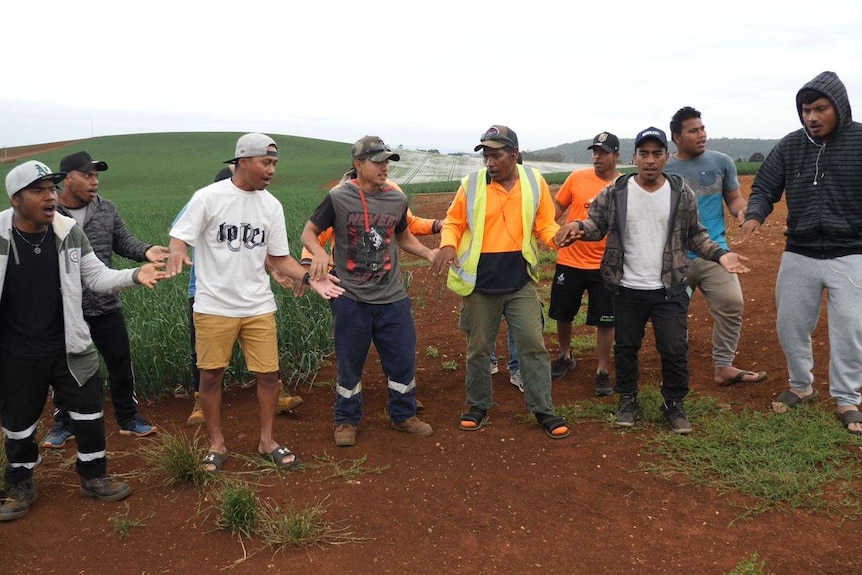 A group of men in casual clothes, some wearing high-vis, stand hand in hand singing in a farm field.
