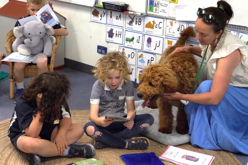 Teacher holding curly brown dog as it looks at small girl sitting on ground reading book., watched by another girl