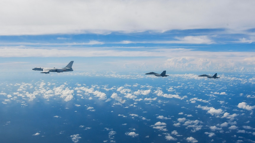 Three military aircraft fly against a bright blue sky dotted with small white fluffy clouds