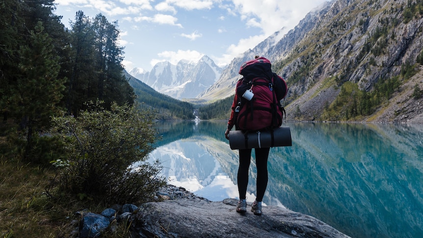 A person wearing a red backpack stands on a rock looking at a lake with their back to the camera.