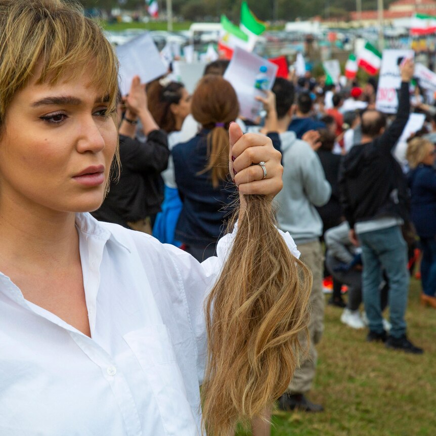 A young woman in a white shirt holds a lock of her hair while crying as protesters march in the background