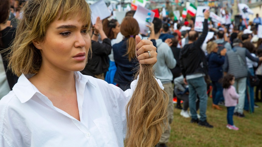 A young woman in a white shirt holds a lock of her hair while crying as protesters march in the background