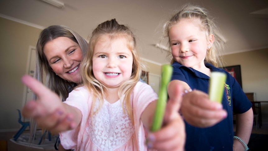 Rebecca Shack with her two young daughters, who are holding out celery sticks to the camera to show off their healthy eating.