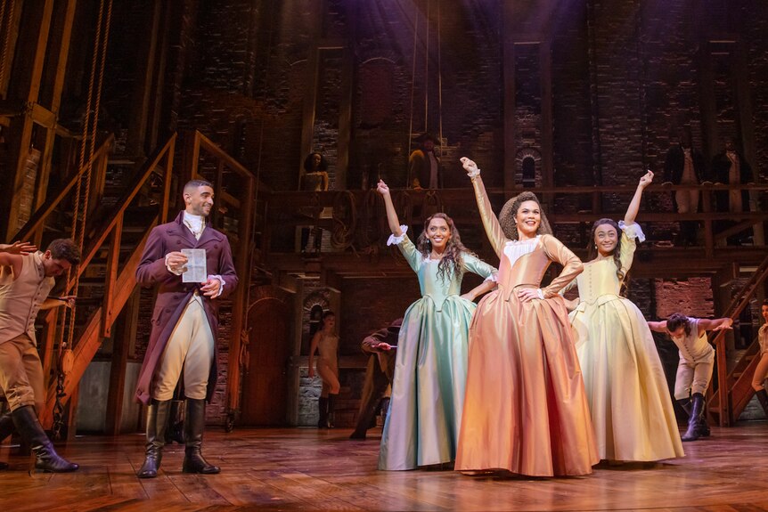 The three Schuyler sisters in 18th century dresses to one side of stage, with Aaron Burr to other side.
