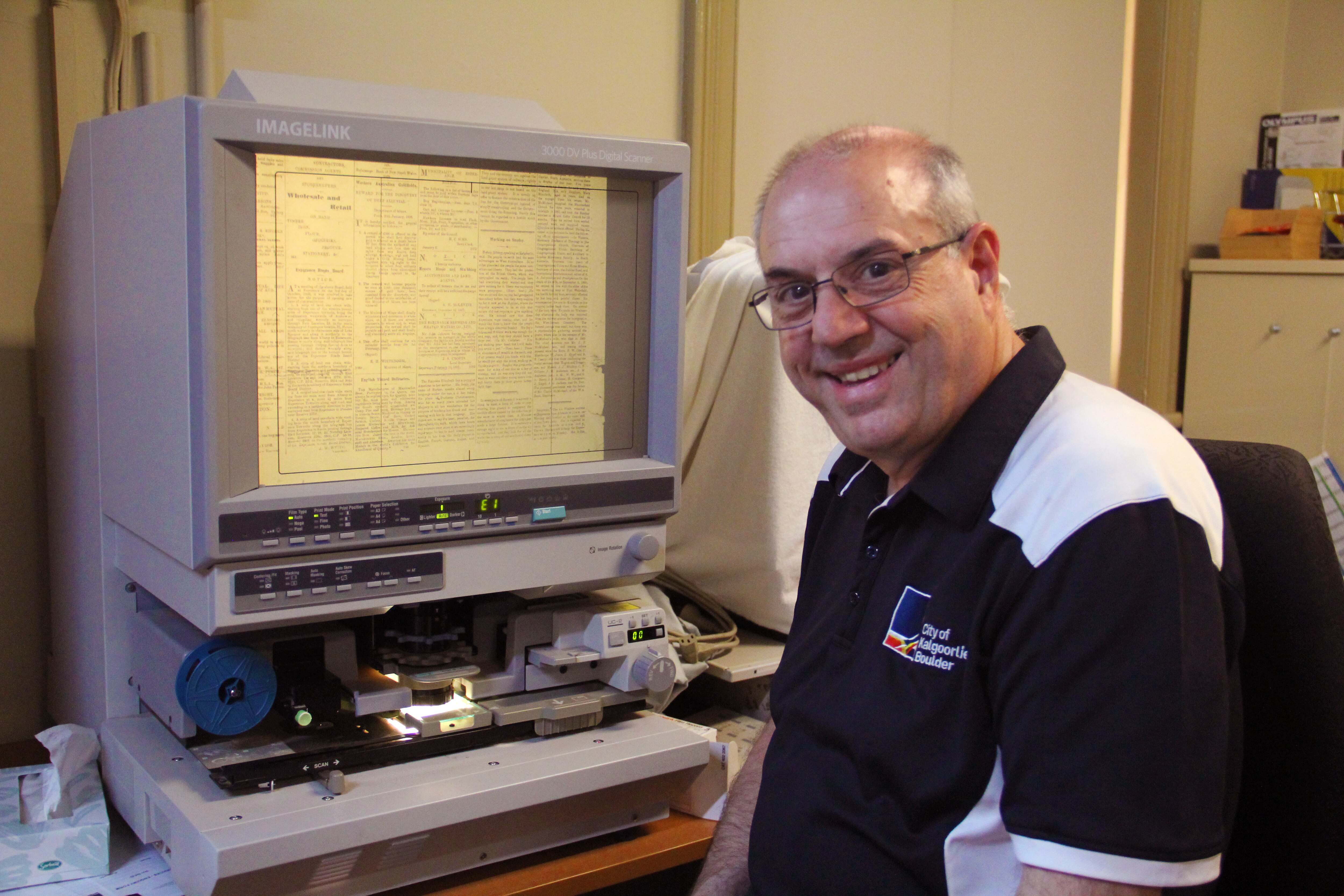 Um homem sorridente de meia-idade está sentado em frente a uma máquina de microfilme.