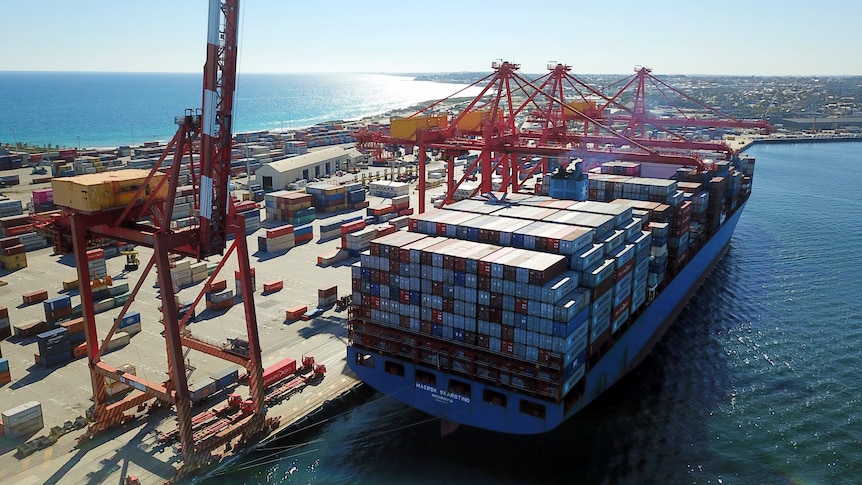 A drone shot of a large container ship alongside a wharf, with cranes and beach in the background.