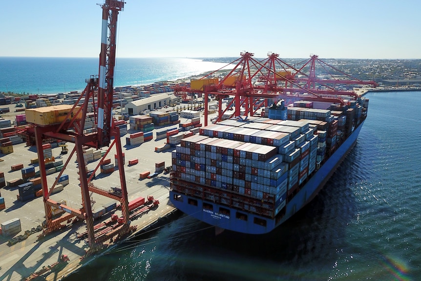 A drone shot of a large container ship alongside a wharf, with cranes and beach in the background.