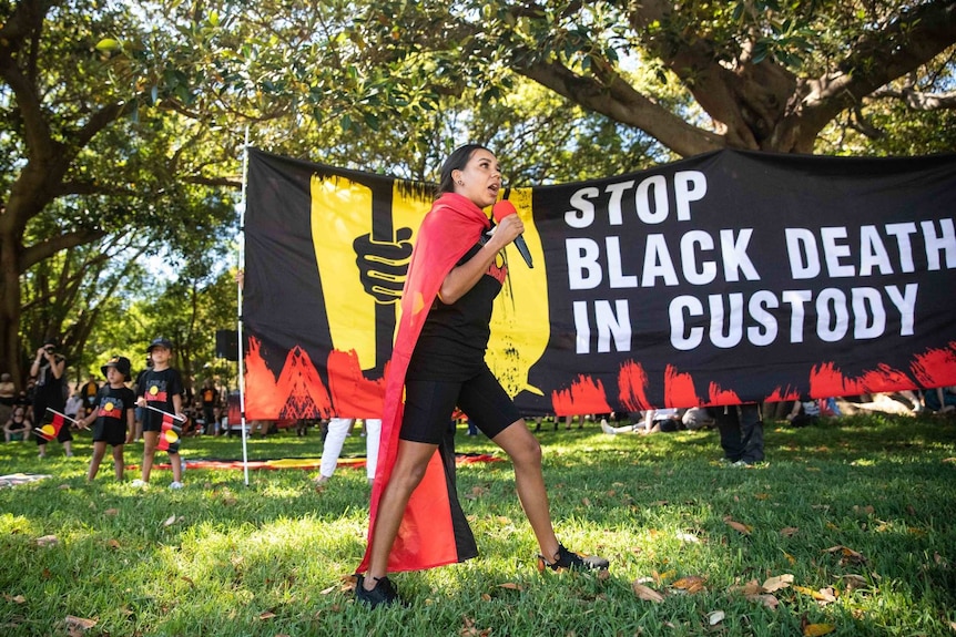 a woman holding a microphone at a park and speaking in front a banner reading stop black deaths in custody