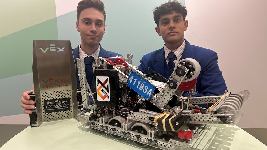 two male students in blue blazers with a robot vehicle and a trophy on the table in front of them
