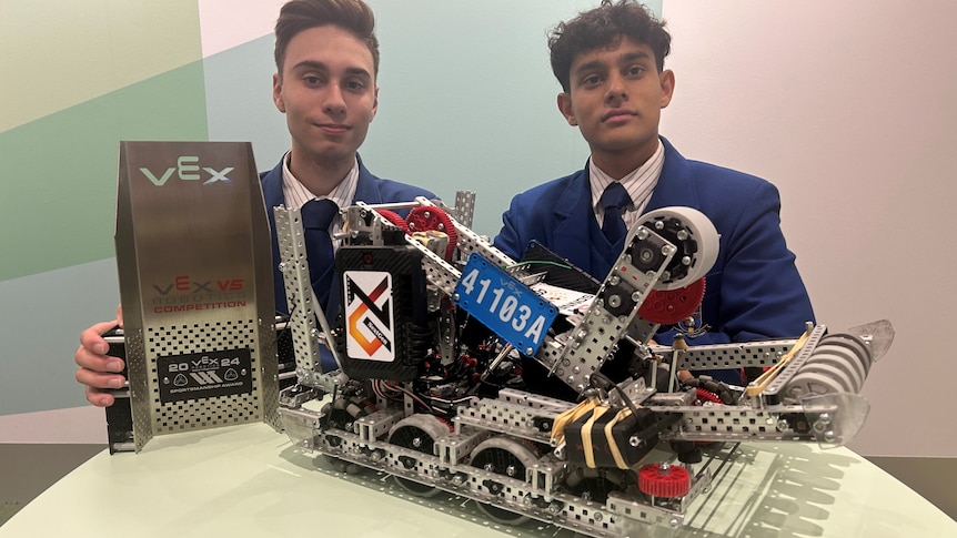 two teenagers in blue school blazers, one on left holding trophy with robot on wheels on table
