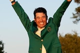 Hideki Matsuyama holds his arms in the air and smiles widely as he wears the green jacket