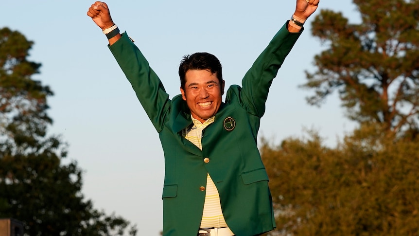 Hideki Matsuyama holds his arms in the air and smiles widely as he wears the green jacket
