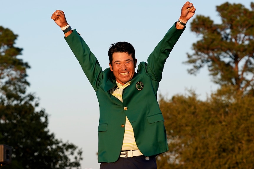 Hideki Matsuyama holds his arms in the air and smiles widely as he wears the green jacket