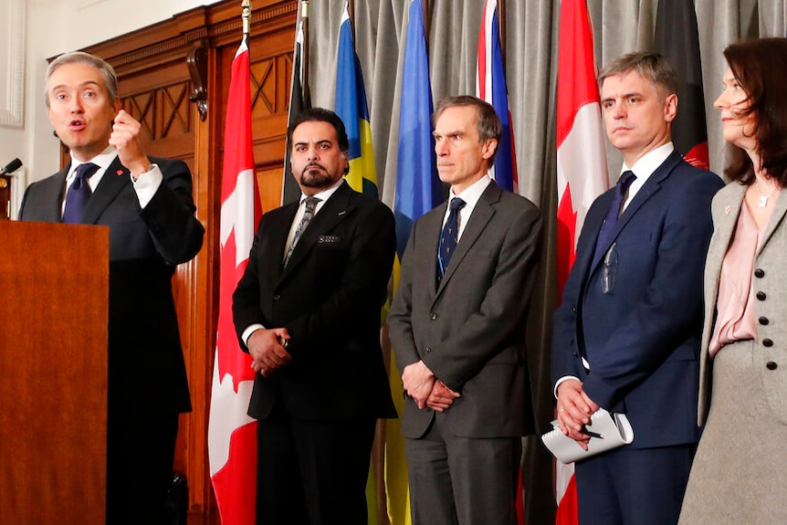 A line of officials wear are in formal workwear as they stand in front of national flags in an ornate office.