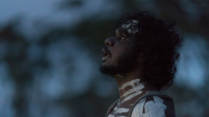 A close-up of a man painted up with ochre looking up to the evening sky.