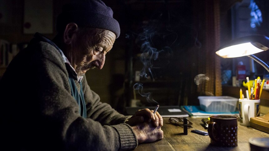 The old orchardist sits at his study desk, eyes downcast, smoke rising from a lit cigarette.