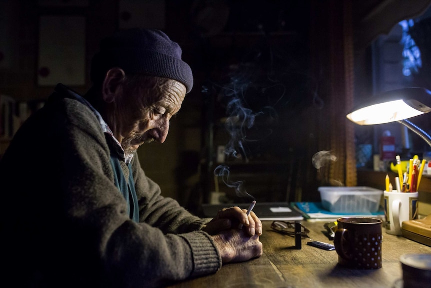 The old orchardist sits at his study desk, eyes downcast, smoke rising from a lit cigarette.