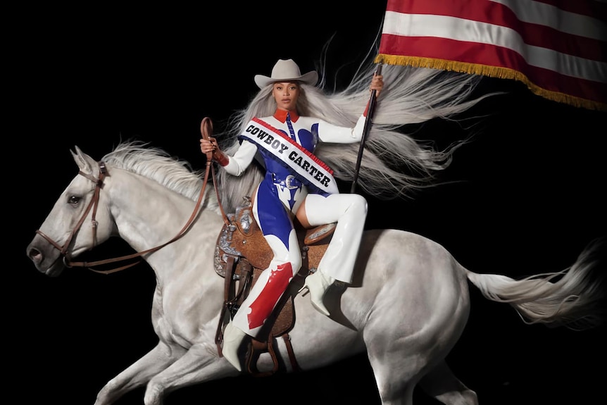 Image of Beyonce sitting on a white horse, wearing a red, white and blue outfit, a cowboy hat and holding an American flag