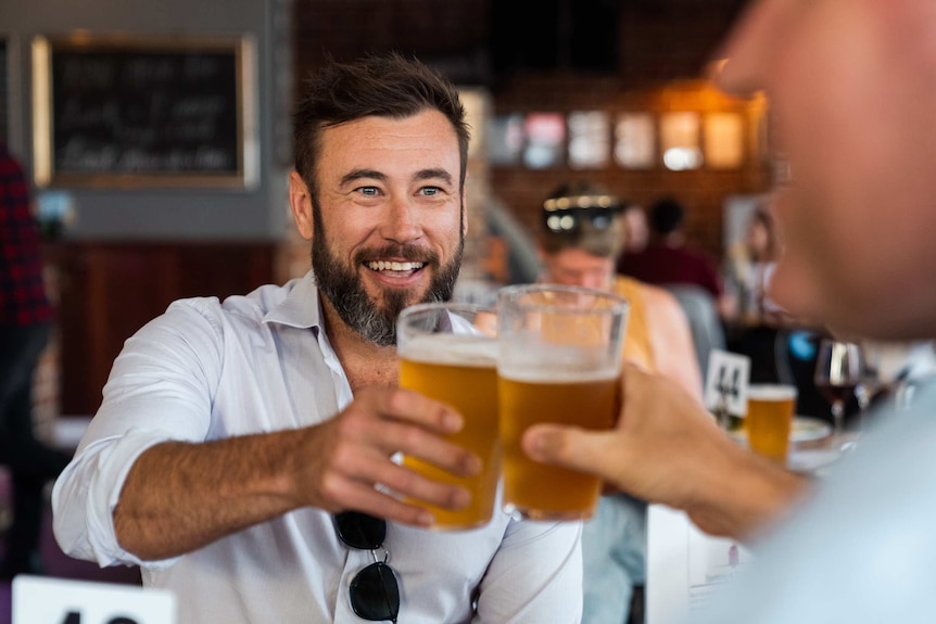 A smiling man holds up a pint of lager in a pub.