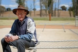 An elderly man sits on a metal bed frame, the base of which is made from chicken wire.