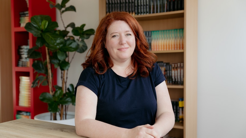 A woman with white skin and red mid-length hair sits a desk with bookshelves and a plant behind her.