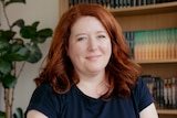 A woman with white skin and red mid-length hair sits a desk with bookshelves and a plant behind her.