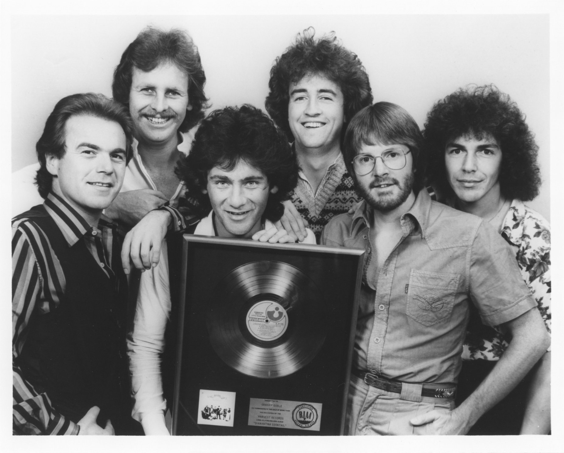 Six 1970s pop musicians crowd together as they smile and hold a gold record