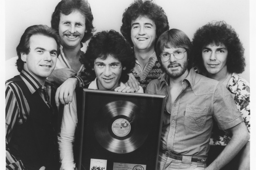 Six 1970s pop musicians crowd together as they smile and hold a gold record