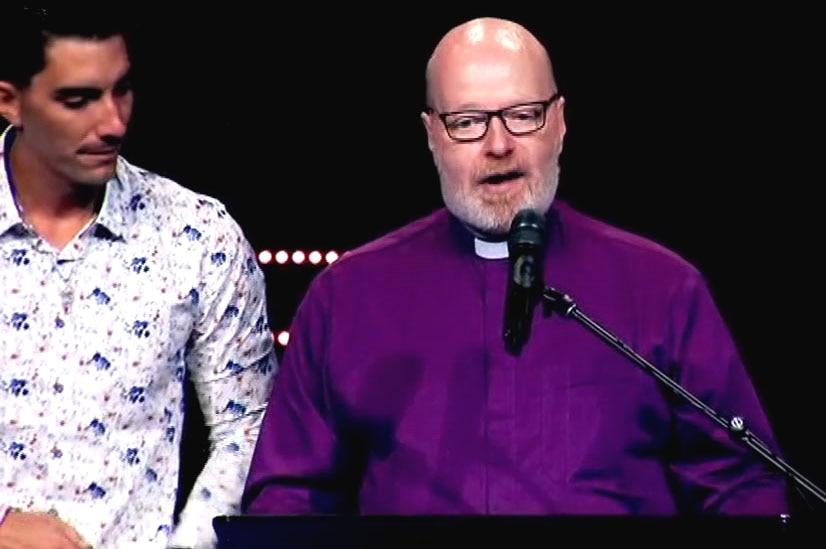 A man in a clerical collar speaking at a lectern, with a younger man beside him