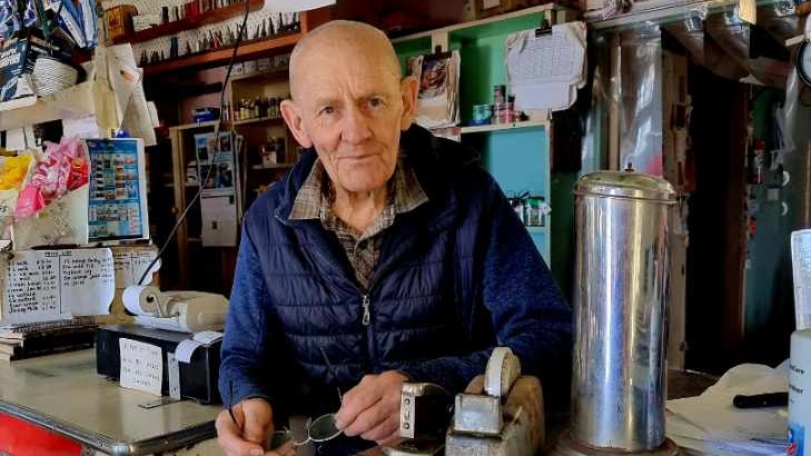 An older man stands behind a shop counter.