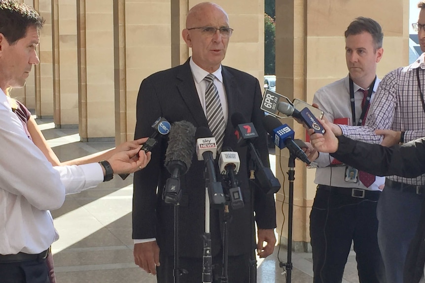 John Quigley standing outside Parliament House speaking to reporters holding microphones.