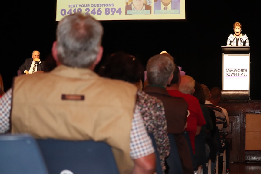 Man and woman on stage talking in background, people sitting in chairs in the foreground.